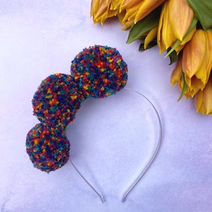 May include: A headband with three colorful pom-poms. The pom-poms are made of yarn in shades of blue, red, yellow, green, and orange. The headband is silver.