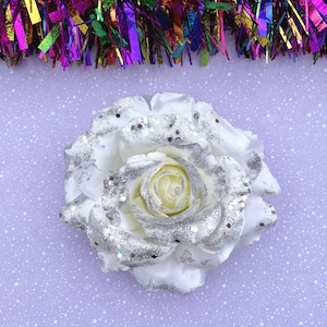 Op de afbeelding: Een grote, witte roos met zilveren glitteraccenten. De roos staat tegen een lichtpaarse achtergrond met witte spikkels. Kleurrijk tinsel staat bovenaan. Bloemenversiering.