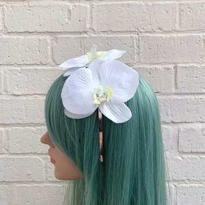 Puede incluir: Pinza para el pelo de orqu&iacute;dea blanca con una barra de metal dorado. La pinza se lleva en una peluca turquesa.
