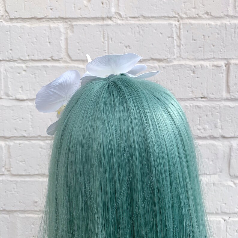 Puede incluir: Una pinza para el pelo de color blanco con dos flores de orqu&iacute;dea blancas. La pinza para el pelo est&aacute; en una peluca de color turquesa.