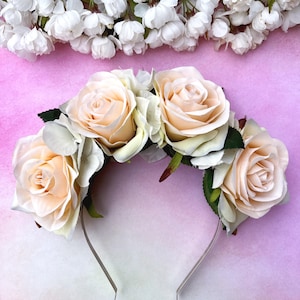May include: A floral crown featuring large peach-colored roses and white accent flowers. The roses are arranged on a silver-colored wire base, with green leaves. The background is a light pink surface with white flowers.