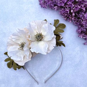 May include: A floral headband featuring two large, off-white fabric flowers with detailed centers and green leaves. The flowers are attached to a thin, silver-toned metal headband, creating an elegant accessory. The background includes purple flowers.