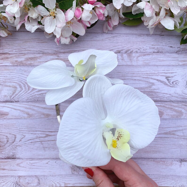 Puede incluir: Dos flores de orqu&iacute;dea blancas con centros amarillos, sostenidas en una mano sobre un fondo de madera clara.