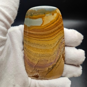 May include: A polished, rectangular stone with a layered pattern of tan, brown, and purple hues. The top of the stone features a light blue and white area. The stone is held by a gloved hand.