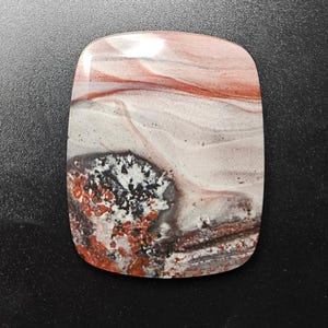 May include: A polished, rectangular gemstone cabochon featuring a smooth surface with swirling patterns of cream, light gray, and deep red. A textured, dark cluster of black and orange mineral inclusions is visible on the lower left side.