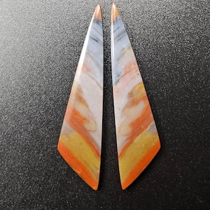 May include: A pair of elongated, triangular stone cabochons featuring a polished surface with swirling bands of orange, yellow, cream, and grey. The stones have a smooth, glossy finish and sharp, pointed tips, displaying a natural, multi-colored pattern.