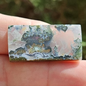 May include: A rectangular polished gemstone featuring a translucent pale pink base with intricate moss-like inclusions in shades of deep forest green, charcoal gray, and creamy white, creating a natural landscape pattern across the stone surface.