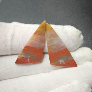May include: Two triangular-shaped polished gemstones featuring horizontal bands of color in shades of vibrant orange, muted gray, and pale yellow. The stones have a smooth, flat surface and are arranged side-by-side with their pointed ends facing upward.