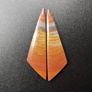 May include: Two polished, triangular-shaped cabochons featuring horizontal bands of orange, yellow, and grey. The stones have a smooth, glossy finish with a pointed top and an angled base, displaying natural mineral layering in warm earth tones.
