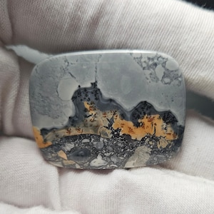 May include: A rectangular, polished stone with a unique pattern. The stone features a blend of gray, black, and orange hues, resembling a landscape. The surface has a smooth, glossy finish, highlighting the natural variations in color and texture.
