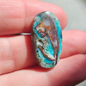May include: An oval-shaped gemstone with a unique pattern of teal, brown, and gold hues. The polished stone displays a marbled effect, with areas of deep teal and lighter turquoise. The gemstone is held in a hand, showcasing its natural beauty.