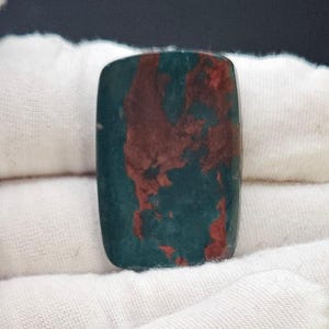 May include: A rectangular gemstone with rounded corners. The stone features a deep teal base color with striking maroon veining and patterns throughout. The gemstone is displayed on a white fabric background.