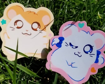 May include: Two cartoon hamster stickers on a green grassy background. One hamster is orange and white with a blue star. The other hamster is white with pink ears and a blue bow.