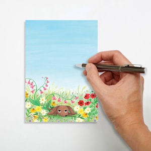May include: A watercolor illustration of a dachshund dog lying in a field of colorful flowers. The dog is brown with big eyes. The background is a light blue sky. A hand holds a pen over the illustration.