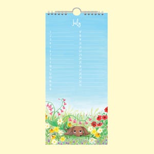 May include: A wall calendar for July, featuring a watercolour sky and a meadow scene with a dachshund. The word "July" is written at the top. Dates are listed on the left and right sides. The calendar is held by a silver metal ring.