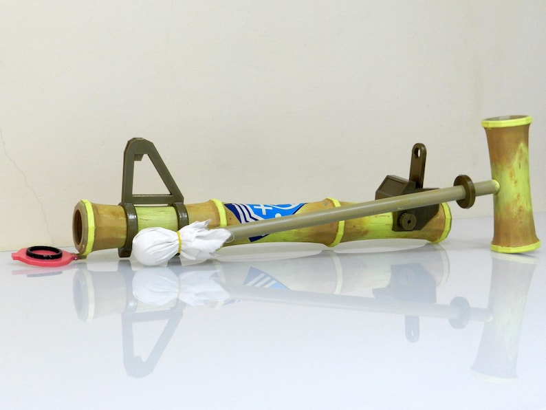 Puede incluir: Una pistola de agua de juguete verde y marr&oacute;n con un pa&ntilde;o blanco sujeto al ca&ntilde;&oacute;n. La pistola tiene un dise&ntilde;o amarillo y azul en el ca&ntilde;&oacute;n. La pistola est&aacute; tumbada sobre una superficie blanca.