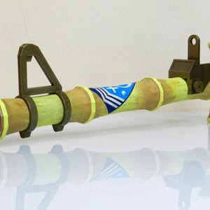 Puede incluir: Un lanzador de cohetes de juguete hecho de madera y pintado de color verde claro. El lanzador de cohetes tiene una tapa rosa en el extremo y un dise&ntilde;o de rayas azules y blancas en el lateral.