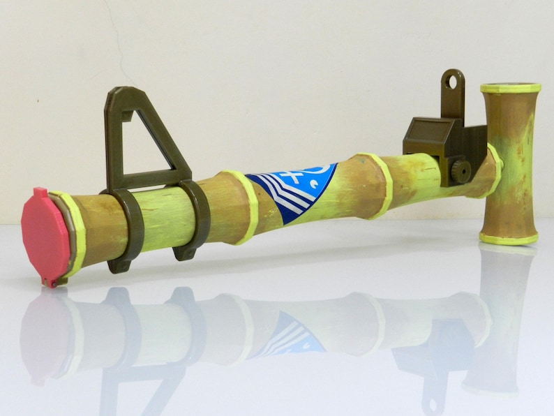 Puede incluir: Un lanzador de cohetes de juguete hecho de madera y pintado de color verde claro. El lanzador de cohetes tiene una tapa rosa en el extremo y un dise&ntilde;o de rayas azules y blancas en el lateral.