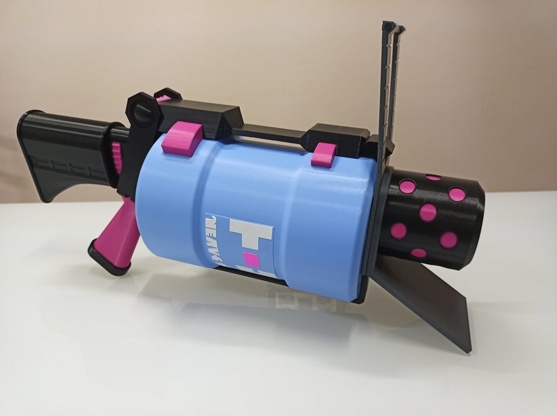 Puede incluir: Una pistola de juguete impresa en 3D con un ca&ntilde;&oacute;n azul, un mango negro y detalles en rosa. El ca&ntilde;&oacute;n tiene un patr&oacute;n de lunares negros y rosas y el texto "HEAVY" impreso en &eacute;l.