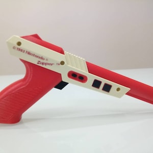 Może przedstawiać: Plastikowa czerwono-biała pistoletowa zabawka NES Zapper. Zabawka ma czarny spust i białą lufę z czarnymi przyciskami. Na boku zabawki widnieje napis "1983 Nintendo Zapper TM".