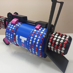 Puede incluir: Un arma de juguete azul y rosa con un mango negro y un dise&ntilde;o tachonado de plata y rojo. El arma tiene la palabra "NEW" impresa en el lateral.