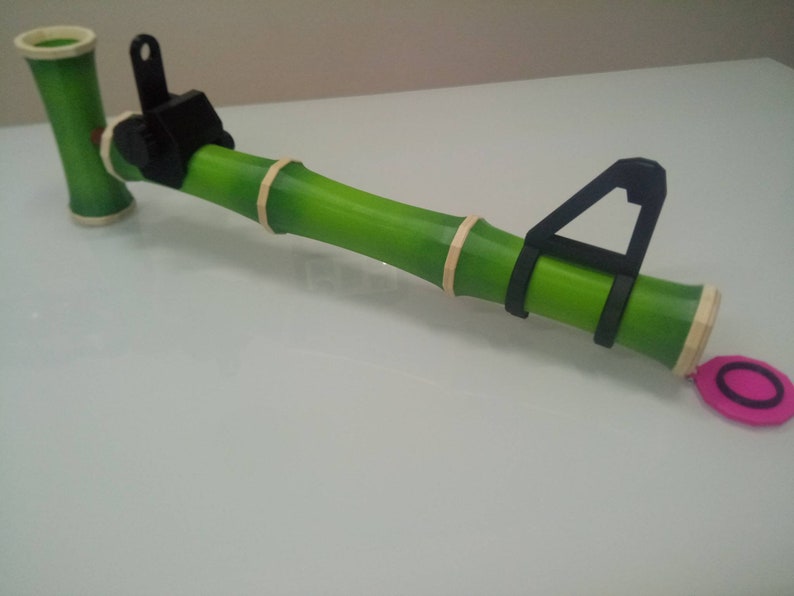 Puede incluir: Un juguete de pistola de bamb&uacute; verde con un gatillo de pl&aacute;stico negro y un mango de pl&aacute;stico negro. La pistola tiene un c&iacute;rculo rosa unido al extremo.