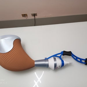 May include: A brown and silver 3D printed object with a blue and black plastic handle. The object is shaped like a horn and has a white tube attached to it.