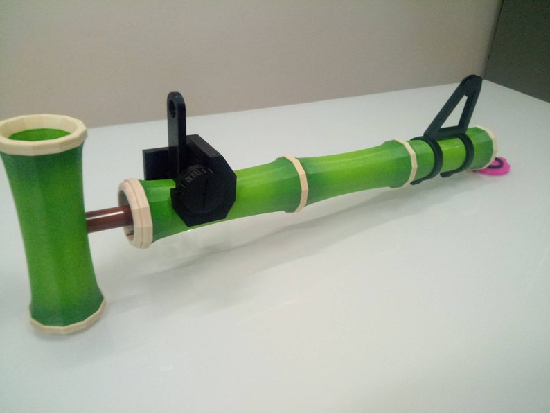 Puede incluir: Un juguete de bamb&uacute; verde con un mango de pl&aacute;stico negro y una clavija de madera marr&oacute;n. El juguete est&aacute; dise&ntilde;ado para ser utilizado como un lanzador de proyectiles.