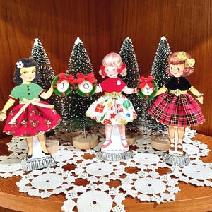 May include: Three vintage paper dolls with Christmas wreaths and miniature Christmas trees. The dolls wear dresses in red, green, and plaid patterns. The dolls are standing on a white lace doily.