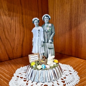 May include: A vintage-inspired Easter figurine featuring two women in dresses, a bunny rabbit, and a sign that reads "Happy Easter". The figurine is displayed on a silver cake pan and a white crocheted doily.
