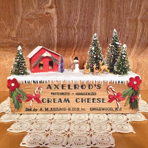 May include: A wooden sign for Axelrod's Cream Cheese with a winter scene. The sign features a red house, snow-covered trees, a snowman, and deer. The sign is decorated with candy canes and red flowers.