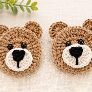 May include: Two circular crochet bear appliqués featuring light brown yarn, small rounded ears, black plastic bead eyes, and a cream-coloured muzzle with a black stitched nose and mouth. These flat, round bear faces are crafted with detailed yarn stitches.