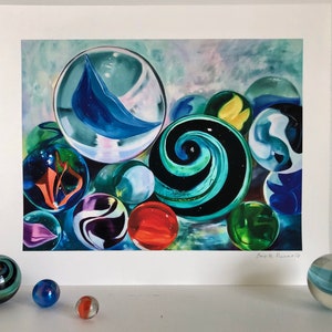 May include: A framed print of a painting depicting a collection of colorful marbles. The marbles are arranged in a cluster, with some overlapping. The background is a soft, abstract watercolor wash in shades of blue and purple. The artist's signature is visible in the lower right corner.