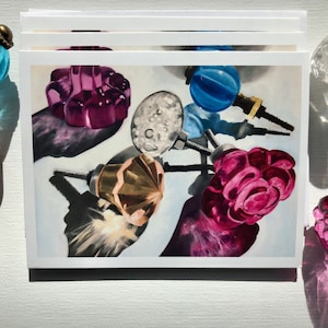 May include: A collection of vintage glass cabinet knobs in various colors and styles, including pink, purple, blue, and gold. The knobs are arranged on a white background and cast shadows.