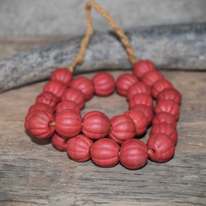 Könnte beinhalten: Eine Halskette aus roten, gerippten Perlen, die auf einer natürlichen Kordel aufgefädelt sind. Die Perlen sind ein tiefes, mattes Rot und haben ein strukturiertes, kürbisartiges Aussehen. Die Halskette wird auf einer verwitterten Holzoberfläche mit einem grauen, strukturierten Objekt im Hintergrund präsentiert.