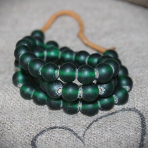 Puede incluir: Una hebra de cuentas de vidrio verde oscuro enhebradas en un cordón marrón. Las cuentas son redondas y tienen un acabado ligeramente esmerilado.