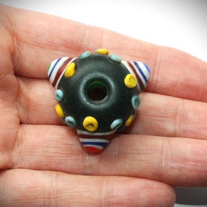 May include: A dark, round glass bead with a central hole and a triangular shape. The bead is decorated with yellow, blue, and red accents. The bead is held in a hand.