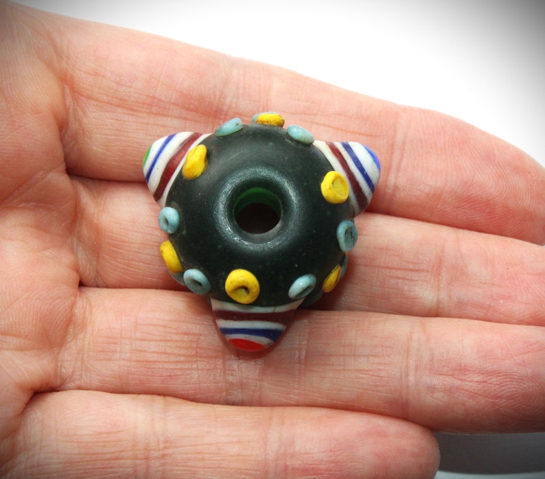 May include: A dark, round glass bead with a central hole and a triangular shape. The bead is decorated with yellow, blue, and red accents. The bead is held in a hand.