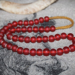 Könnte beinhalten: Eine Halskette aus roten Glasperlen mit einer braunen Schnur. Die Perlen sind matt und haben eine leicht unregelmäßige Form.