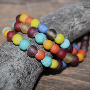 Könnte beinhalten: Nahaufnahme von drei Armbändern mit Perlen in verschiedenen Farben, darunter Orange, Gelb, Blau und Türkis. Die runden Perlen scheinen aus Glas oder einem ähnlichen Material zu bestehen. Die Armbänder liegen auf einem Stück verwittertem Holz.