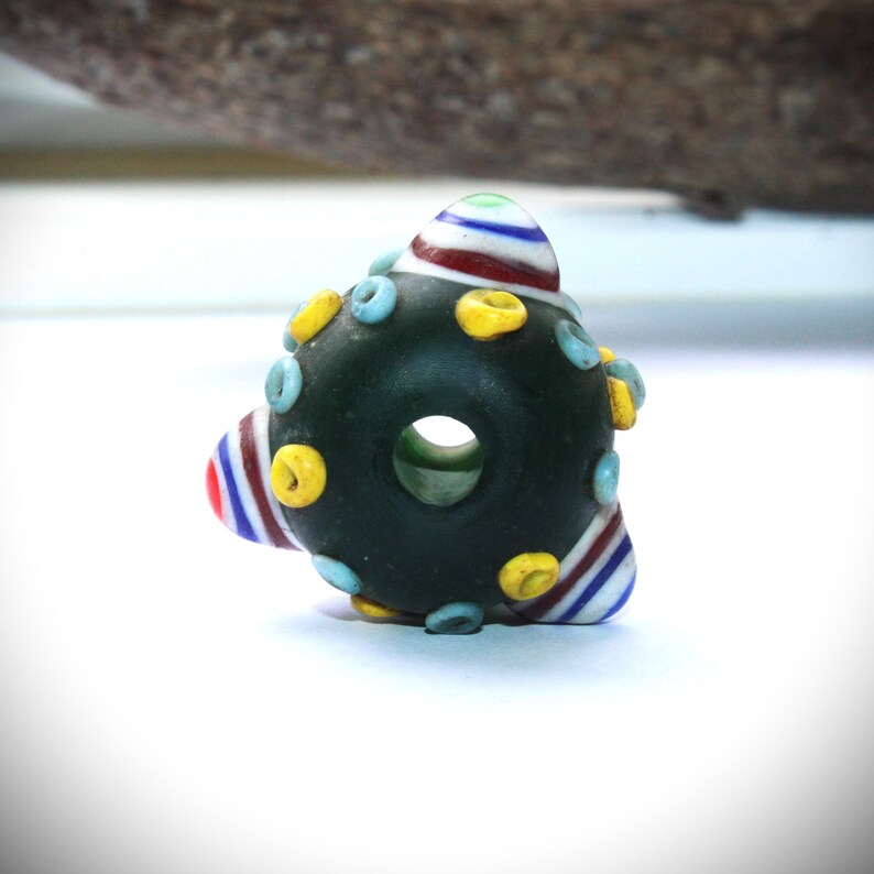 May include: A dark green glass bead with a large central hole. The bead is decorated with colorful accents, including blue, yellow, red, and white stripes and dots. The bead has a unique, organic shape.