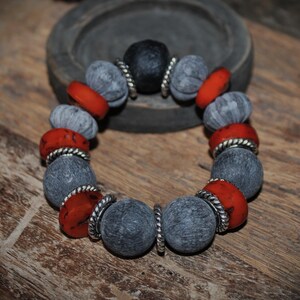 May include: A handmade bracelet featuring large, gray, and red beads. The bracelet's beads are interspersed with silver-colored spacers. The bracelet is displayed on a wooden surface, showcasing its earthy tones and textures.