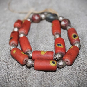Könnte beinhalten: Eine Halskette mit roten, zylindrischen Perlen, die mit gelben und blauen Augenmotiven verziert sind. Die Perlen sind durch silberfarbene Metallabstandshalter getrennt. Die Halskette wird auf einem neutralfarbenen Stoff gezeigt.