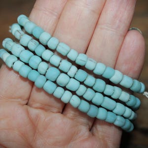 Könnte beinhalten: Eine Strähne aus hellblauen Glasperlen mit leicht mattierter Oberfläche ist auf einem weißen Faden aufgereiht. Die Perlen sind klein und rund und in einem lockeren, fließenden Muster angeordnet.