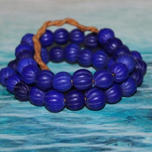 Könnte beinhalten: Ein Strang tiefblauer, geriffelter Perlen, aufgereiht auf einem beigefarbenen Band, liegt auf einer blau-türkisfarbenen Oberfläche. Die Perlen haben eine matte Oberfläche und sind gleichmäßig angeordnet.