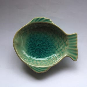 Può includere: Un piatto in ceramica a forma di pesce nei toni del verde. Il piatto ha una superficie strutturata e screpolata e una forma curva. Le pinne e la coda sono dettagliate con linee a coste. Il piatto viene probabilmente utilizzato per servire cibo o come oggetto decorativo.