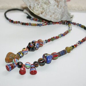 May include: A vibrant beaded necklace featuring a mix of glass beads in red, blue, yellow, and black. The necklace includes round, teardrop, and cylindrical beads strung on a black cord. The beads display diverse patterns.