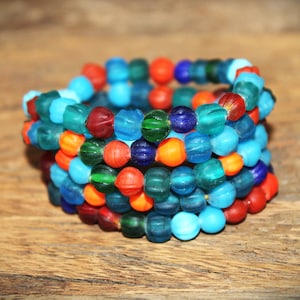 Op de afbeelding: Een stapel kleurrijke glazen kralen armbanden in tinten blauw, groen, oranje en rood. De armbanden zijn gemaakt van kleine, ronde kralen met een getextureerd oppervlak.