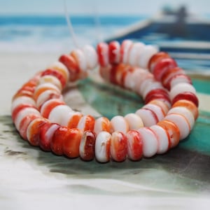 Könnte beinhalten: Eine Kette aus orangefarbenen, roten und weißen Muschelkugeln. Die Perlen sind aneinandergereiht und bilden eine kreisförmige Form. Der Hintergrund zeigt ein verschwommenes Bild des Meeres und eines Bootes.