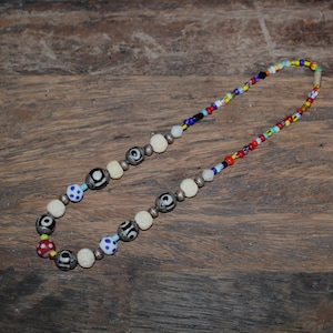 Könnte beinhalten: Eine Halskette mit einer Vielzahl von bunten Perlen. Die Halskette hat weiße, schwarze, blaue, rote und gelbe Perlen. Die Perlen sind mit einem silberfarbenen Metall aufgefädelt. Die Halskette wird auf einer Holzoberfläche präsentiert.